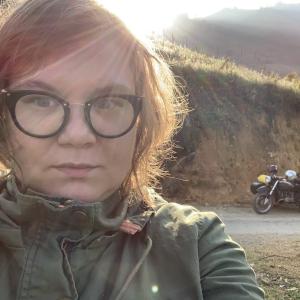 Bree is backlit by a glorious sun setting over the mountains of Ha Giang, Vietnam. A vintage soviet Ural motorcycle rests placidly in the background, awaiting for the journey to resume.